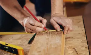 Ręce mierzą drewnianą kantówkę czerwoną kredką i miarką. To pierwszy krok, jak zrobić huśtawkę ogrodową z kantówek.