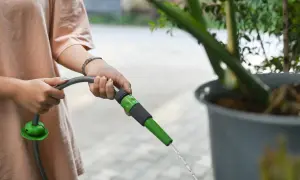 Dłoń trzymająca wąż ogrodowy, z którego strumień wody zasila dużą donicę z rośliną.