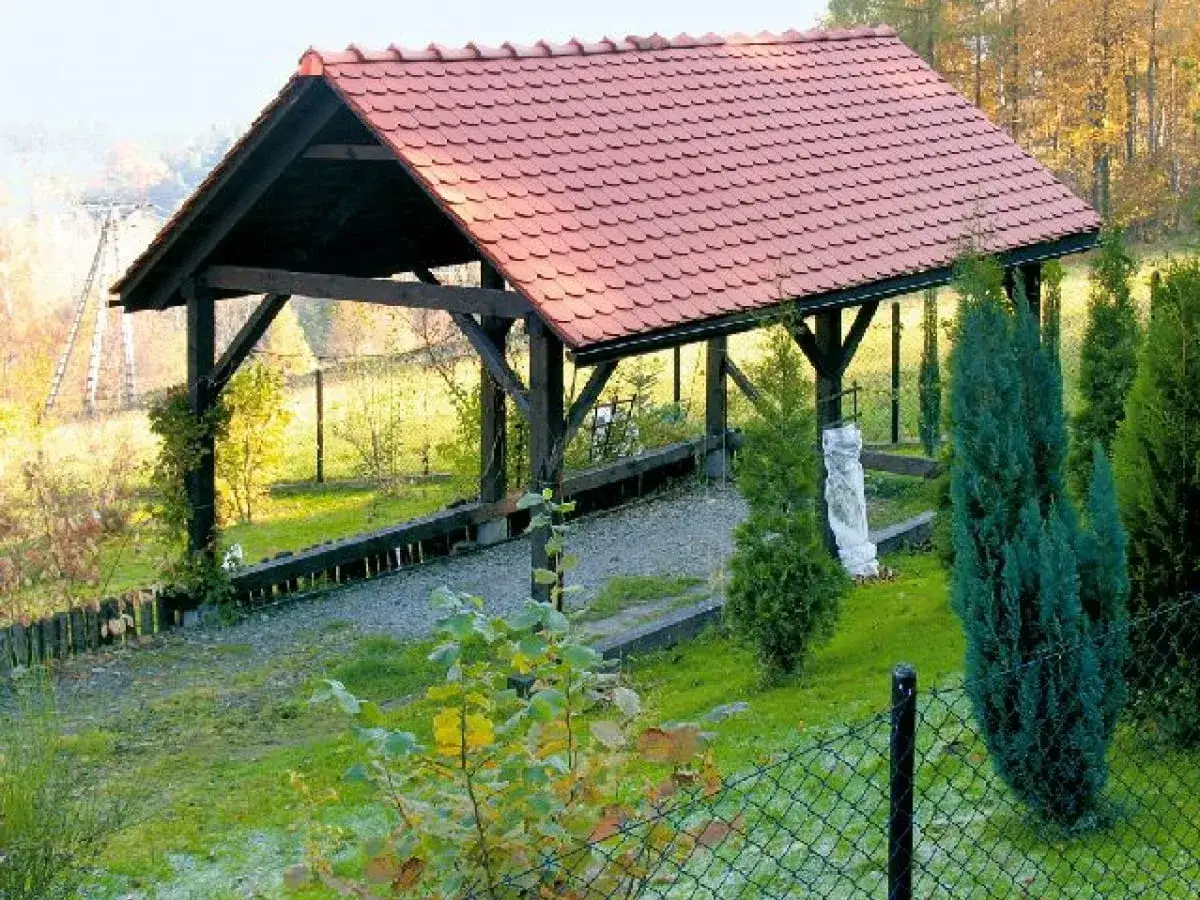Drewniana wiata na słupach betonowych z czerwonym dachem, otoczona zielenią i jesiennym krajobrazem.