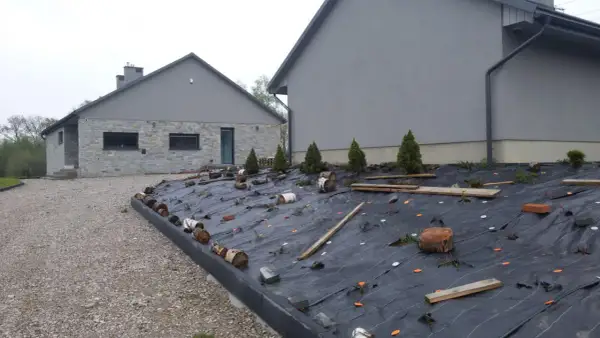 Jak położyć agrowłókninę na skarpie: czarna agrowłóknina zabezpiecza skarpę przed erozją, z widocznymi pachołkami, drewnem i roślinami.